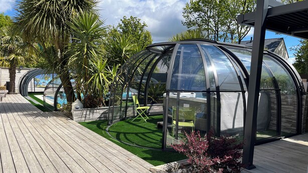 Un espacio de estar al aire libre con paredes de vidrio, un piso de madera y abundante vegetación tropical, bajo un cielo despejado.