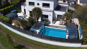 Una casa moderna de dos pisos con piscina, rodeada de vegetación y otras viviendas.