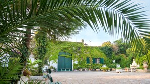 A courtyard with a stone floor, a green bench, and lush greenery including palm leaves. - Apartment with Pool and Park in Villa Oasi (Santa Barbara)