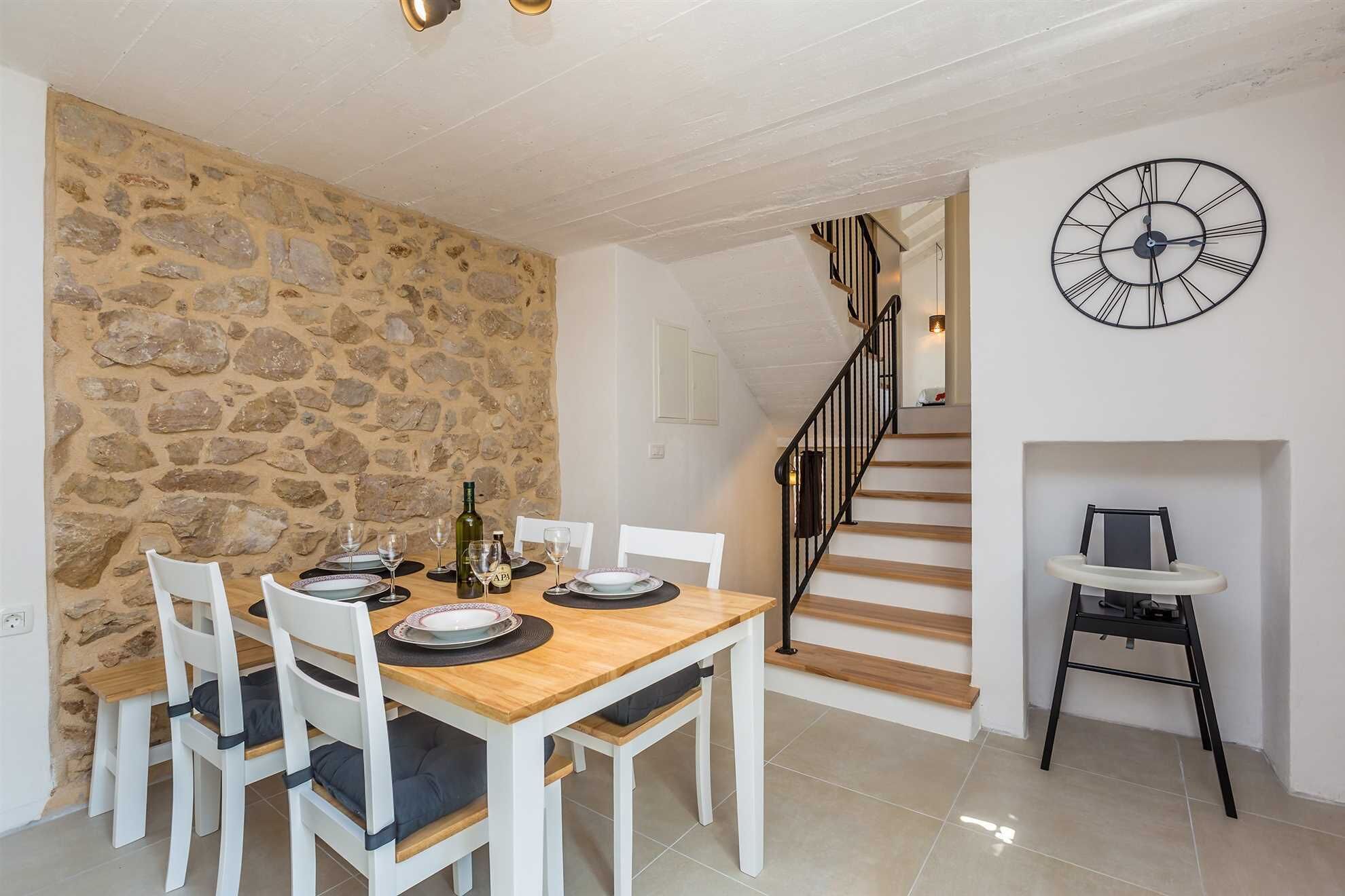 A dining area with a stone wall, a wooden table, white chairs, and a black clock on the wall.