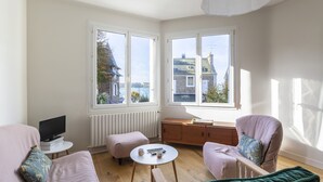 A room with a sofa, armchair, and ottoman, a small table with a remote control, and a view of buildings through large windows. - Le Timonier - Two Bedroom Apartment, Sleeps 4 (Saint-Malo)