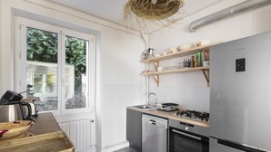 A modern kitchen with a stainless steel refrigerator, a dishwasher, and a gas stove. There are wooden shelves above the sink and a window with a view of greenery outside. - Le Timonier - Two Bedroom Apartment, Sleeps 4 (Saint-Malo)