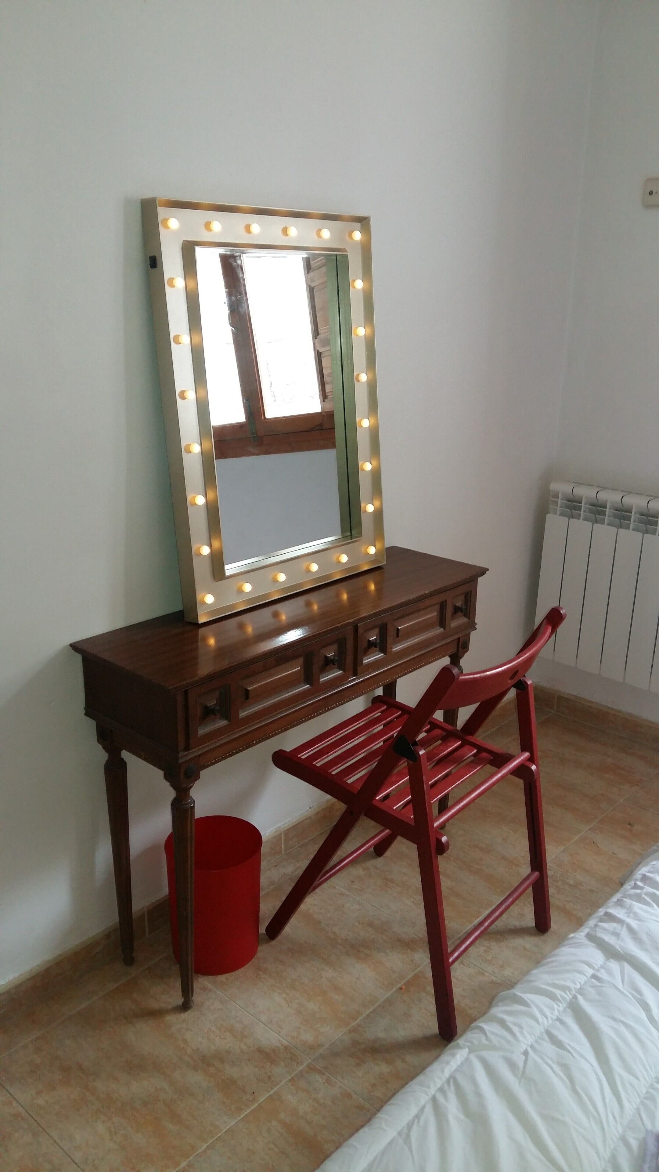 Une pièce comprenant une coiffeuse, un miroir avec éclairage, une chaise rouge, une poubelle rouge et un radiateur.