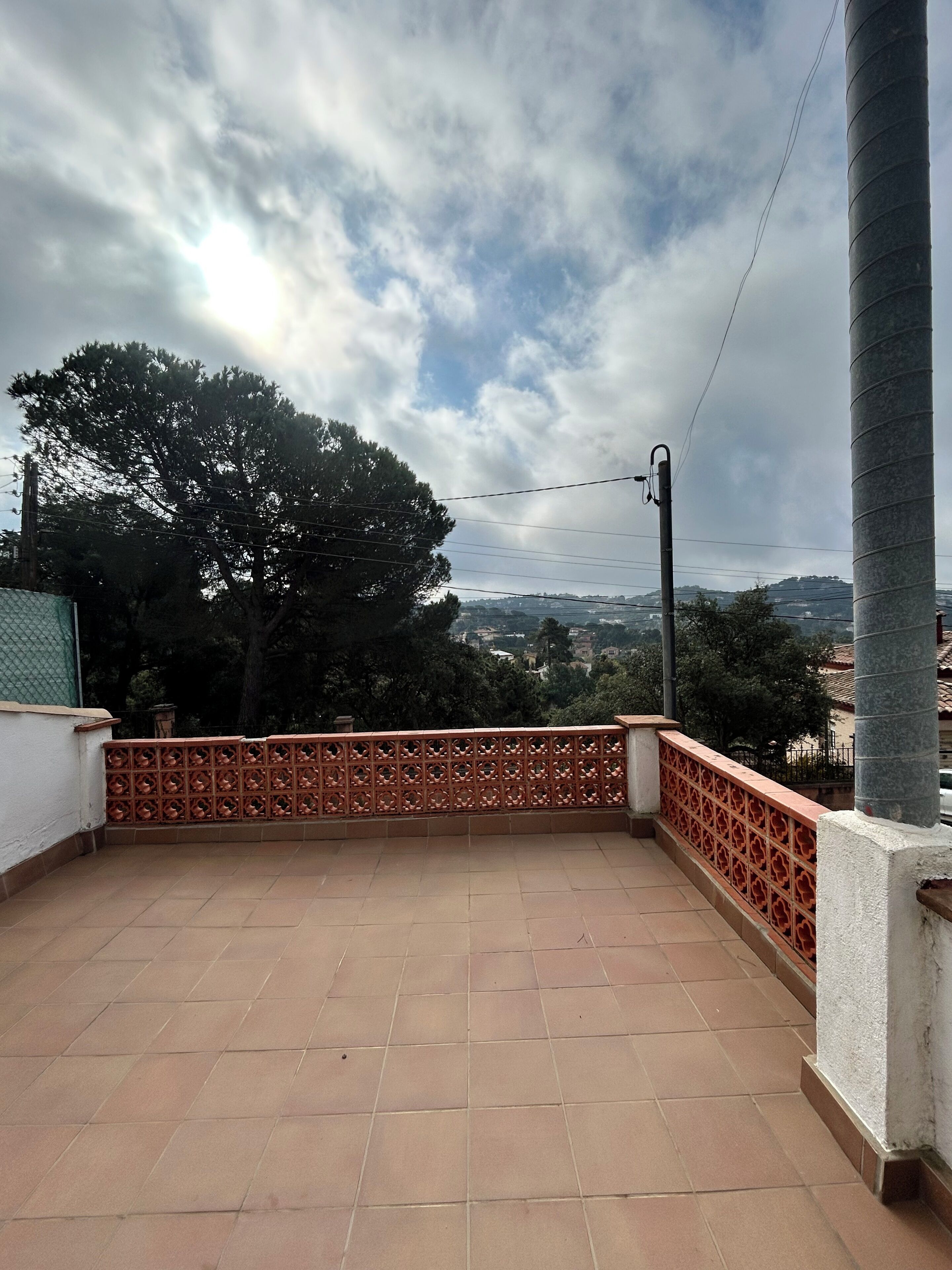 Une terrasse dotée d’un sol carrelé, d’une clôture décorative et offrant une vue sur un arbre et des bâtiments éloignés, le tout sous un ciel nuageux.