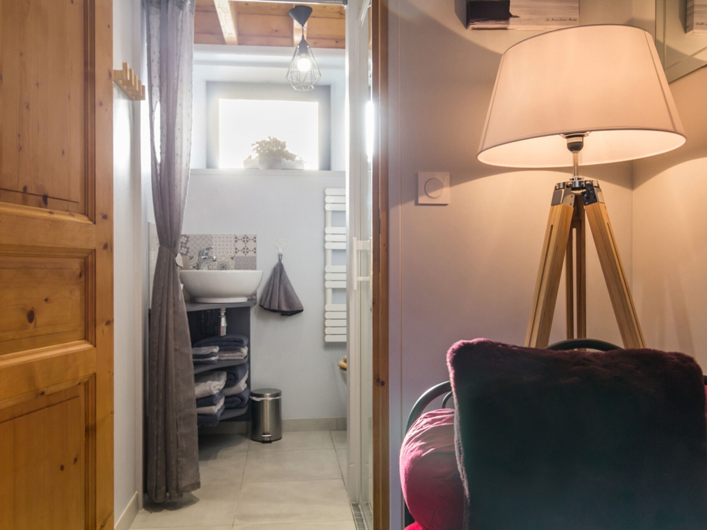 Un couloir étroit mène à une salle de bain équipée d’un lavabo, d’un porte-serviettes et d’une petite poubelle. Une lampe et une chaise sont visibles au premier plan.