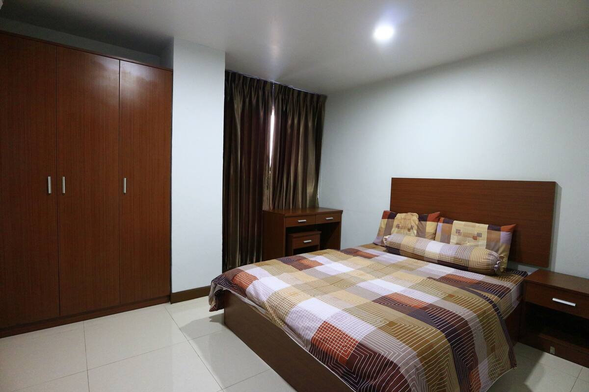 A bedroom with a large wardrobe, a bed with a patterned bedspread, a nightstand, and a bedside table.
