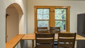 Uma área de refeições com mesa e cadeiras de madeira, uma janela com vista para vegetação e um candeeiro fixado na parede.