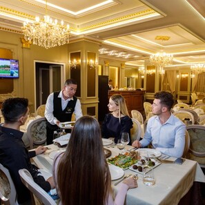 A dining area with a chandelier, a waiter serving, and a television on the wall. - Elite Palace (Tirane)