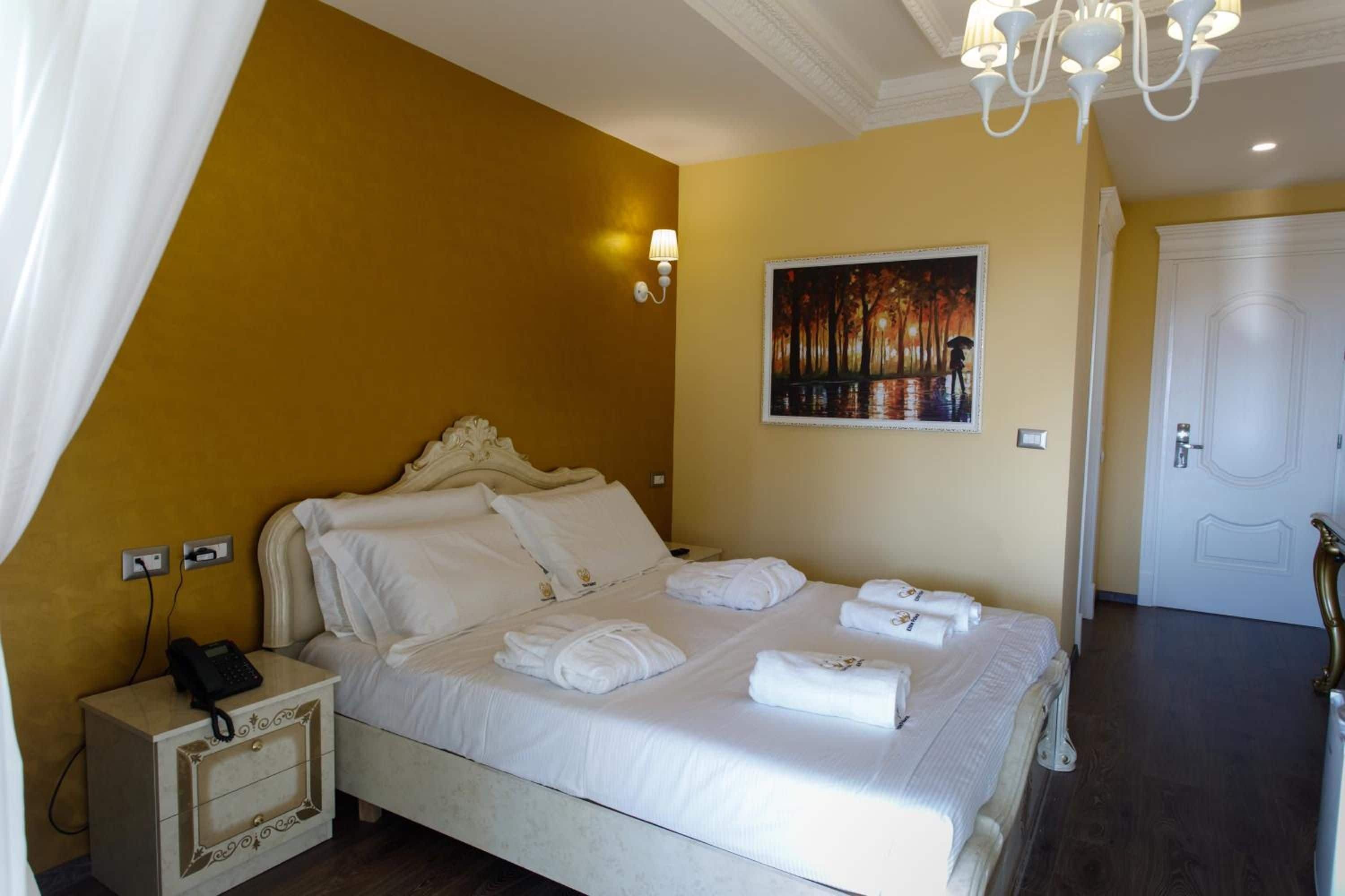 A hotel room with a bed, a painting on the wall, a white door, and a chandelier.