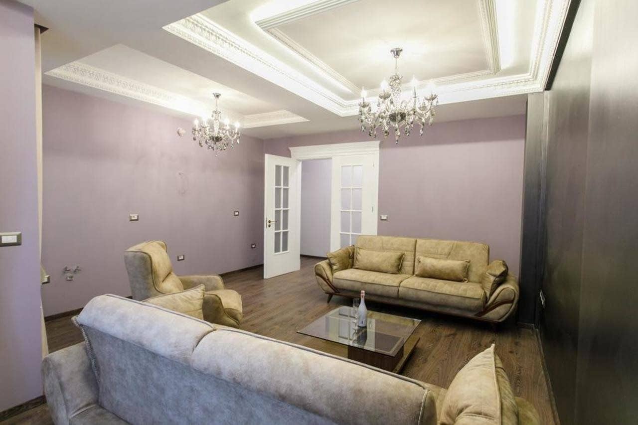 A living room with a beige sofa, two armchairs, a glass coffee table, and a chandelier.