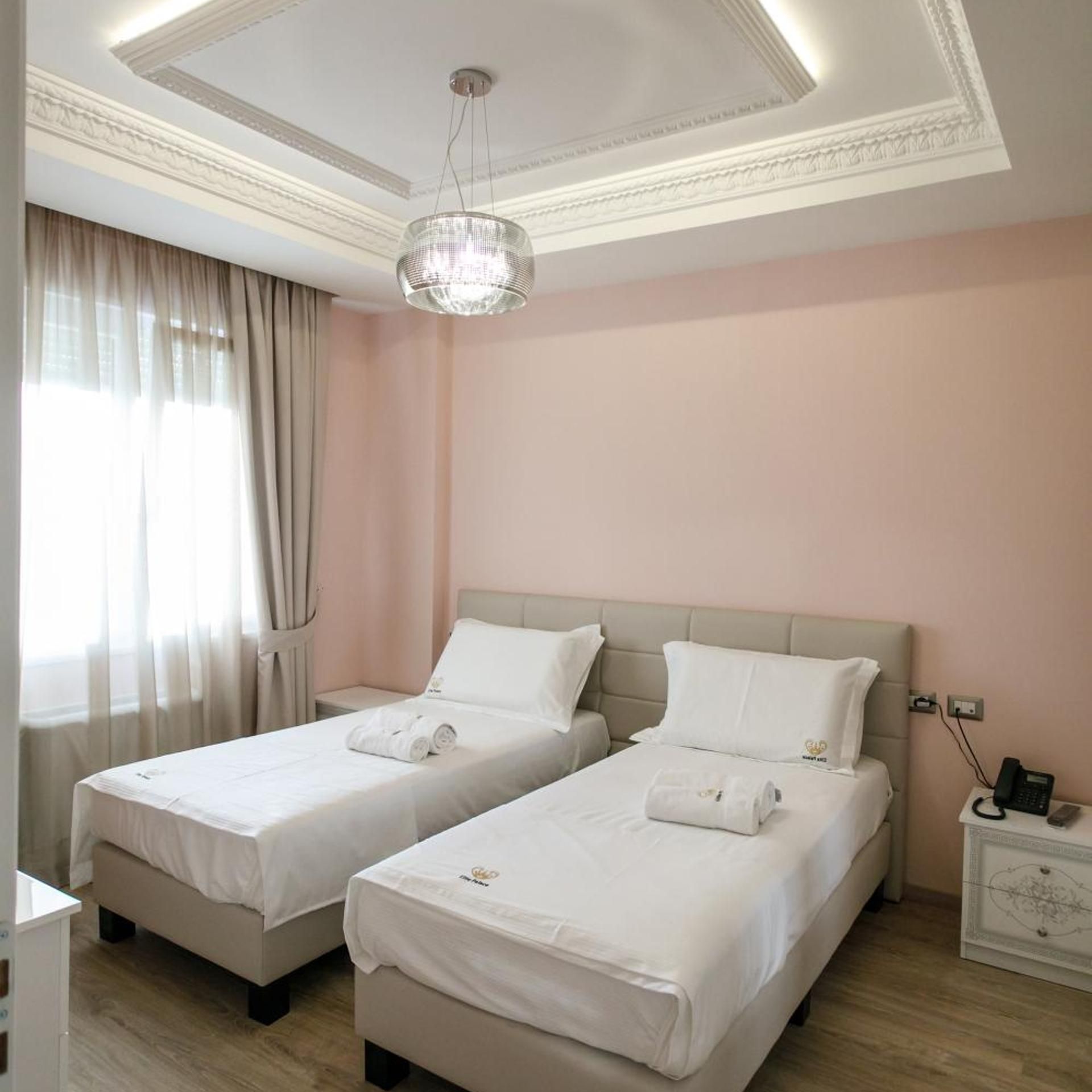 A hotel room with two beds, a chandelier, a nightstand, and a window with curtains.