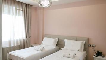A hotel room with two beds, a chandelier, a nightstand, and a window with curtains.