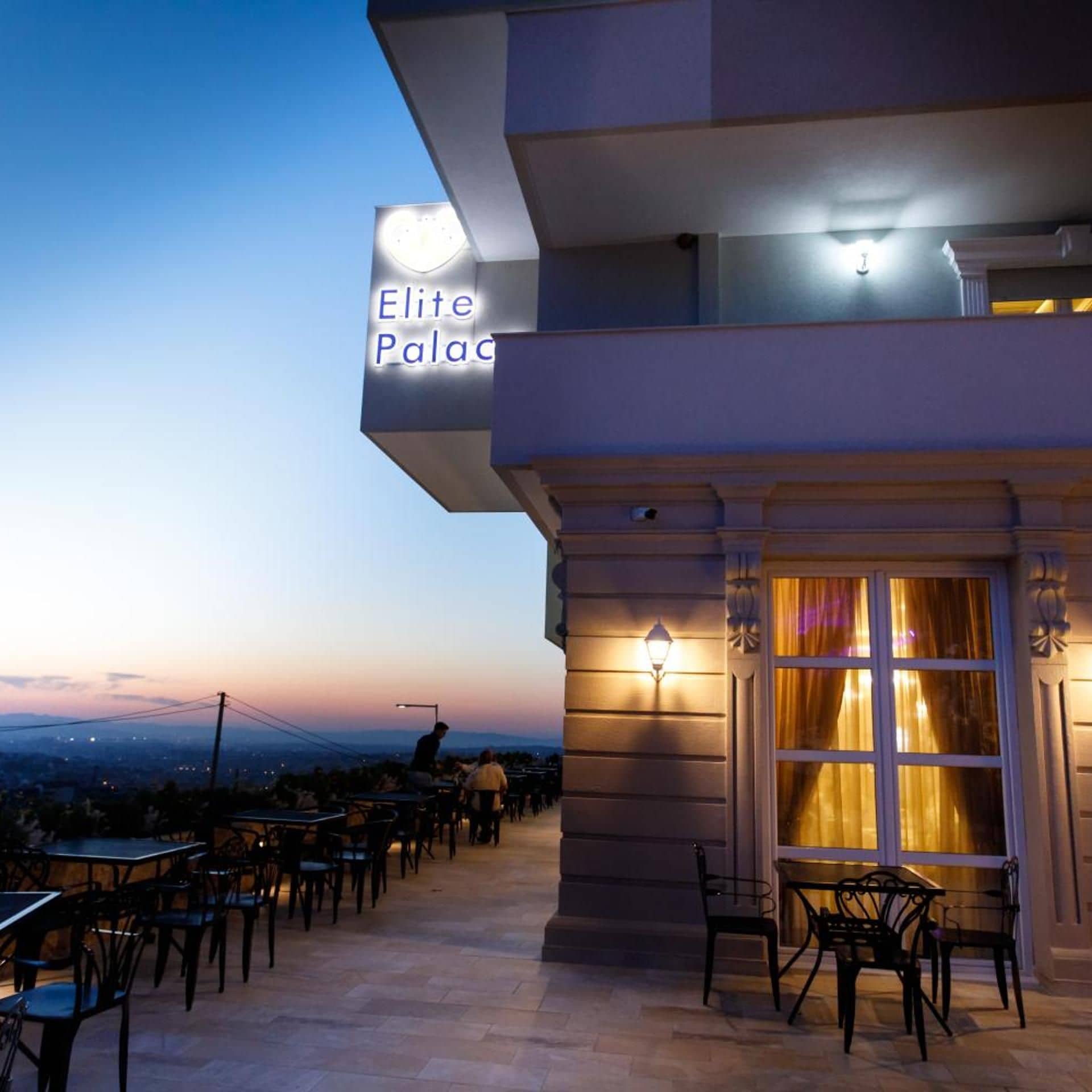 Zona salotto all'aperto con tavoli e sedie, un cartello che riporta 'Elite Palace' e vista del paesaggio sullo sfondo.