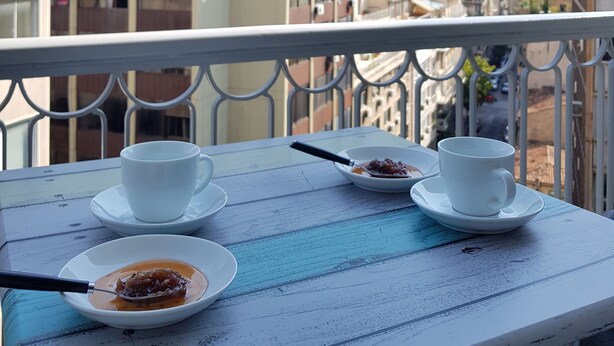 Un balcón con una mesa dispuesta para dos personas, que incluye una taza blanca, un platillo y un cuenco con comida. El balcón ofrece vistas a un paisaje urbano con edificios y un cielo despejado.