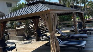 A wooden deck with lounge chairs and a covered area, surrounded by palm trees and a building in the background.