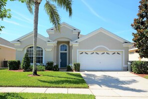 A single-story house with a white garage door, a front entrance with a blue door, and a small front yard with a palm tree and shrubs. - The Explorer's Club - South Pool/Spa, Gated Area! (Kissimmee)