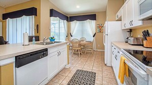 Una cocina funcional con armarios blancos, lavavajillas, microondas y tostadora. Hay una mesa de comedor con sillas y una ventana con cortinas.