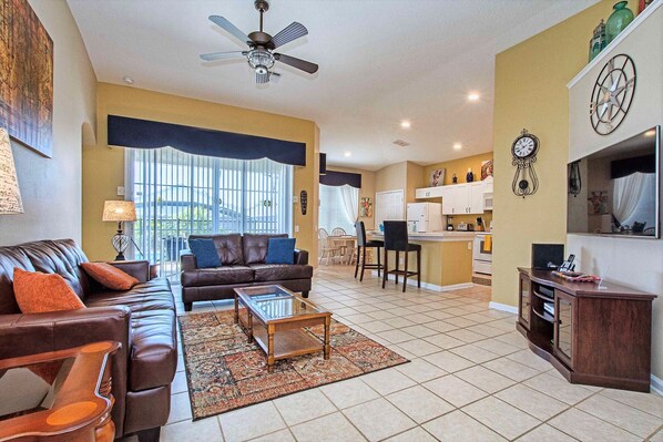 A living room with a brown leather sofa, a glass coffee table, and a patterned rug. There is a ceiling fan, a kitchen area with cabinets, and a dining table with chairs. - The Explorer's Club - South Pool/Spa, Gated Area! (Kissimmee)