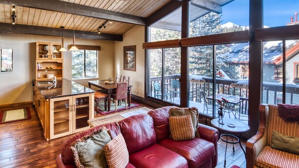 Interior acogedor de una cabaña con techo de madera, un sofá de cuero rojo, zona de comedor y ventanas amplias con vistas a un paisaje nevado.