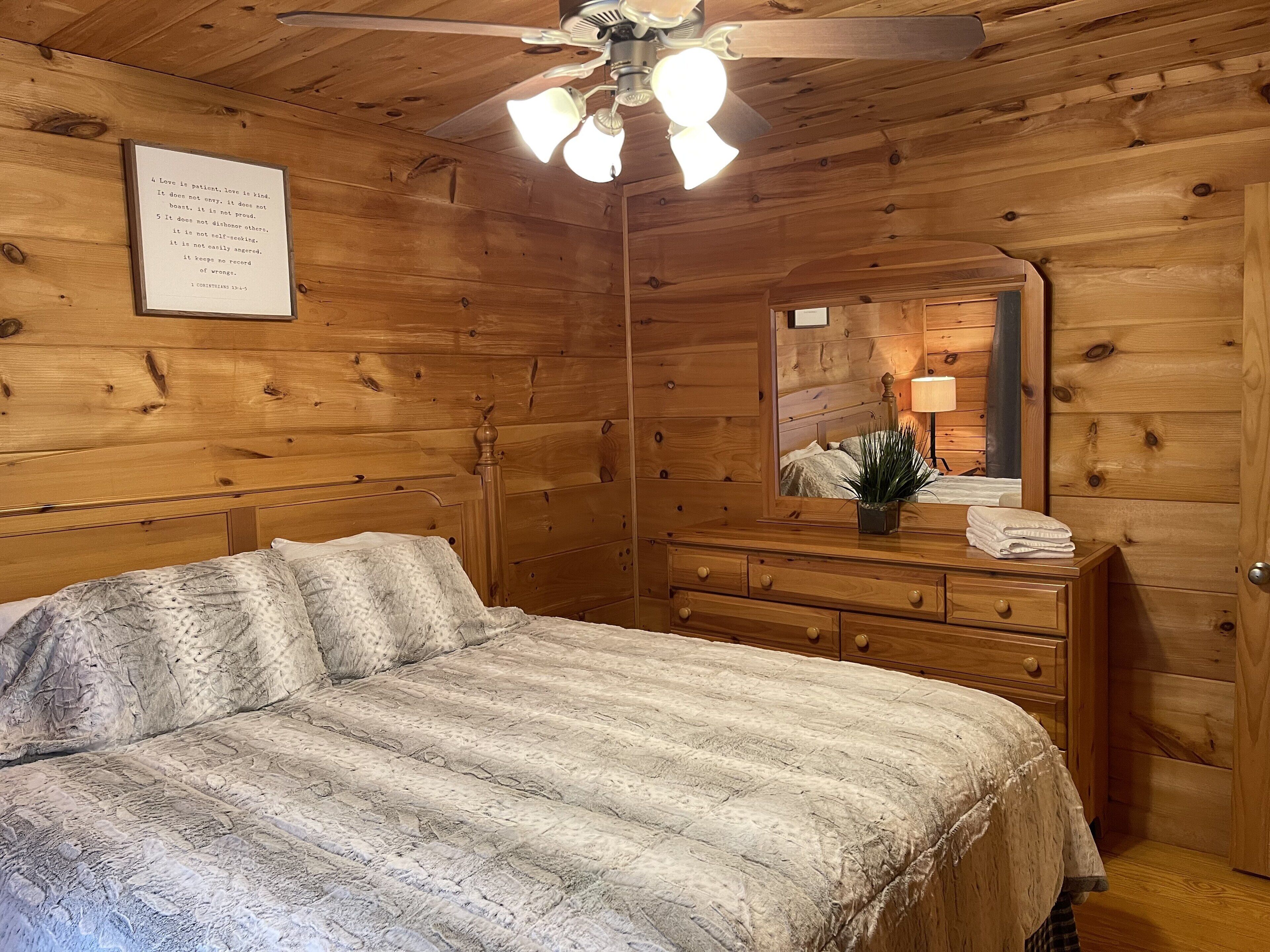 Une chambre dans une cabane en bois, avec un lit, une commode, un miroir, un ventilateur de plafond et une citation encadrée accrochée au mur.