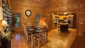 A wooden cabin interior with a dining area, kitchen, and a clock on the wall.