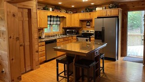 A wooden cabin kitchen with a central island, wooden cabinets, a refrigerator, and a window with a plaid curtain.