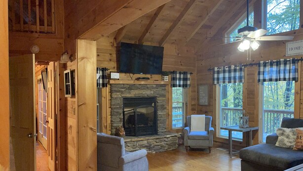 A cozy cabin interior with a stone fireplace, wooden floors, and a large window overlooking a forest.
