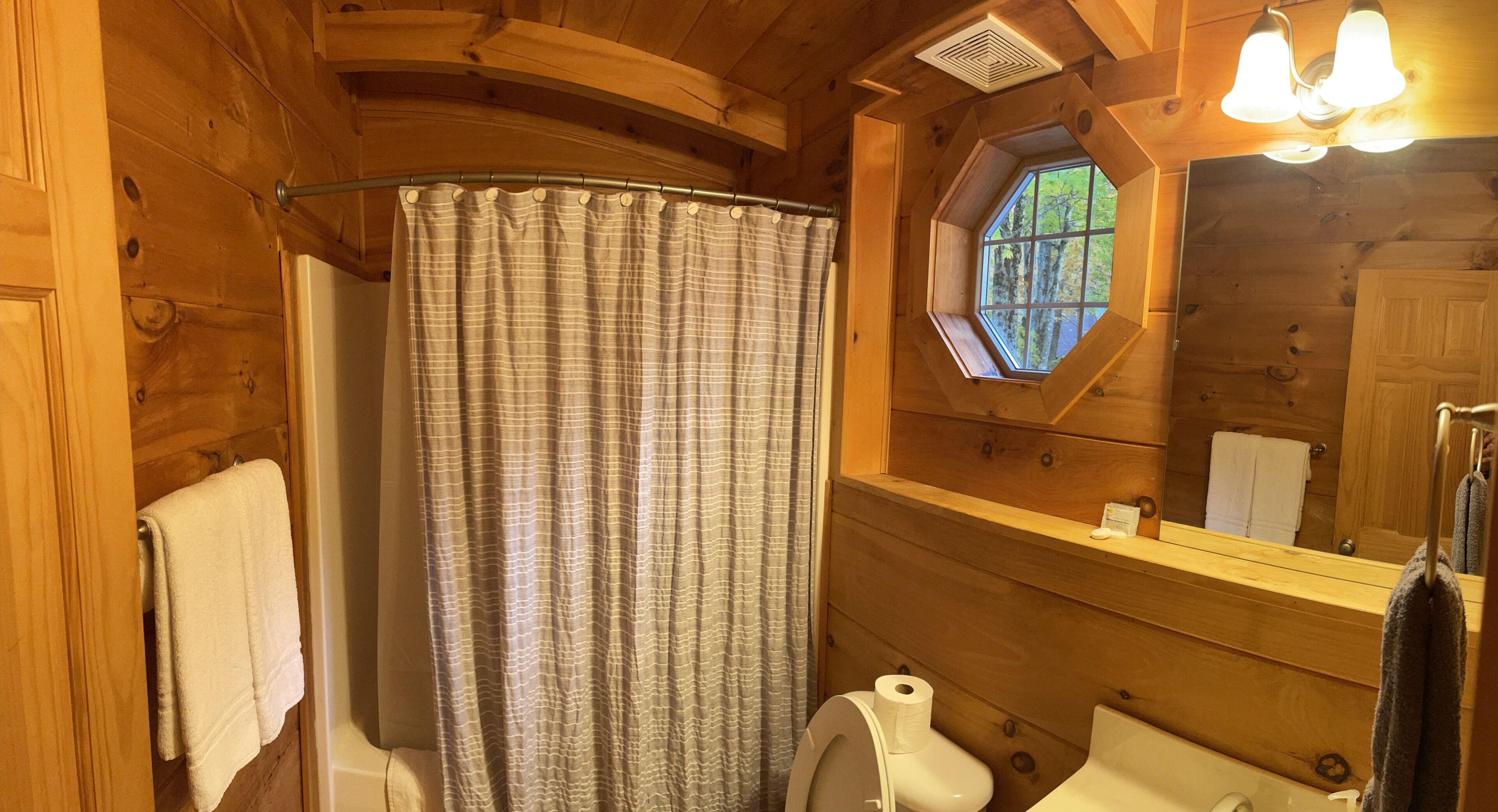 Une salle de bain rustique, aménagée dans une cabane en bois, avec un rideau de douche, des toilettes, un lavabo et une fenêtre donnant sur la forêt.