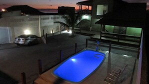 A swimming pool at night with a blue light, a car parked nearby, and a house in the background. - CASA DE FRENTE PARA A PRAIA DE FIGUEIRA COM PISCINA - ARRAIAL DO CABO (Arraial do Cabo)