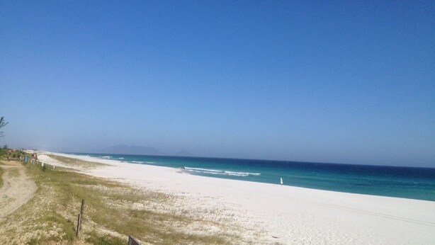 A beach with a clear sky, sandy shore, and a few people in the distance. - CASA DE FRENTE PARA A PRAIA DE FIGUEIRA COM PISCINA - ARRAIAL DO CABO (Arraial do Cabo)