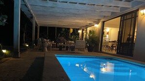 Una piscina exterior con un porche cubierto, asientos y plantas en macetas.