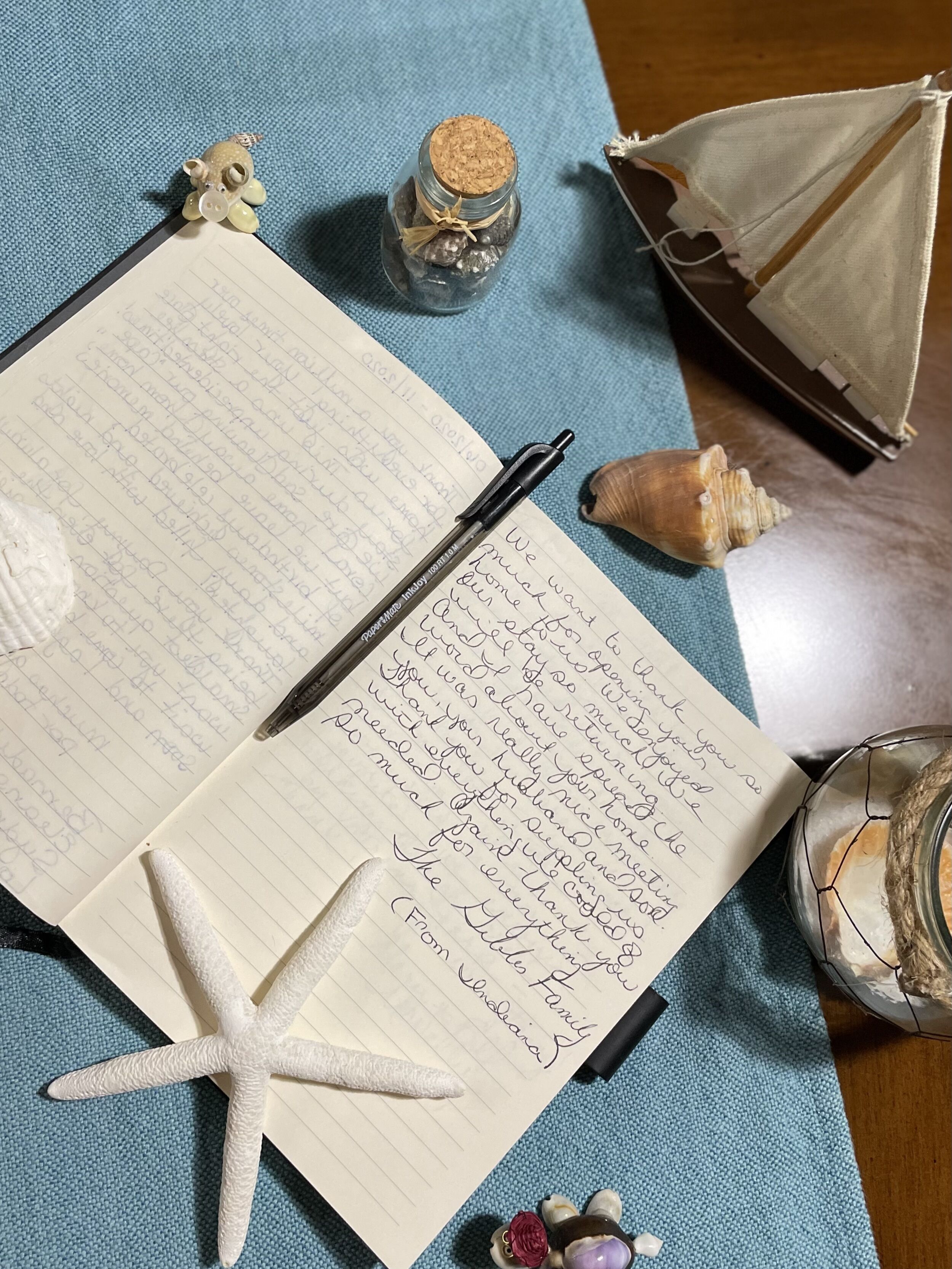An open notebook with handwritten notes, a pen, a starfish, seashells, and a small jar with a cork lid on a blue fabric surface.