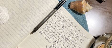 An open notebook with handwritten notes, a pen, a starfish, seashells, and a small jar with a cork lid on a blue fabric surface.