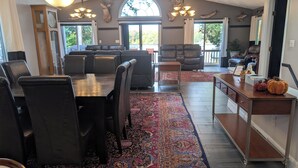 A spacious dining area with a large table and chairs, a patterned rug, and a view of the outdoors through a large window. - Amazing Lake House With All Amenities (Remsen)