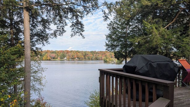 A wooden deck overlooking a lake with trees and houses in the background. - Amazing Lake House With All Amenities (Remsen)