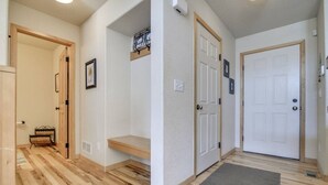 Un couloir doté d’un plancher en bois, de murs blancs et d’un banc. On y trouve trois portes, dont une avec un paillasson, ainsi qu’une petite étagère avec des photos.