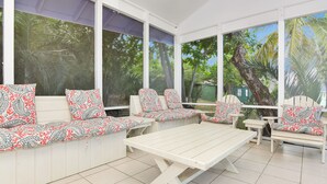A covered patio with white furniture, patterned cushions, and large windows overlooking a tropical garden. - Plumbago - Paradise in the Bahamas with Ocean views (George Town)