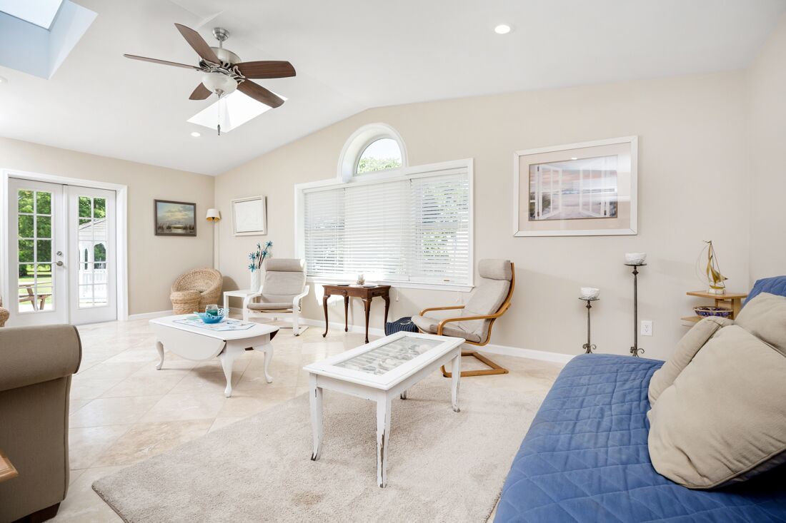 A spacious living room with a ceiling fan, a large window with blinds, a blue bed, and a white coffee table.