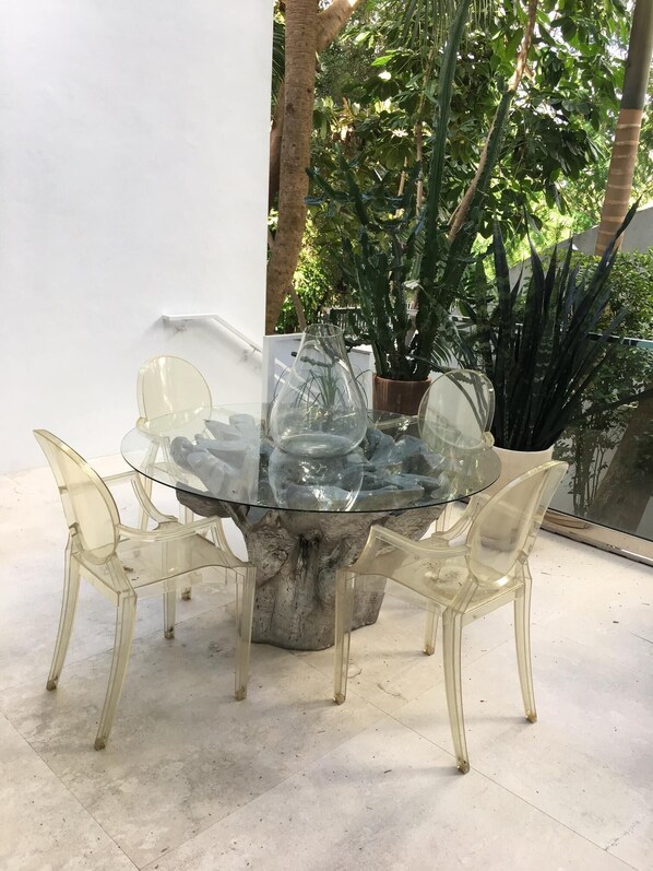 Une table à plateau en verre, entourée de quatre chaises transparentes et de plantes en pot, dans un décor extérieur.