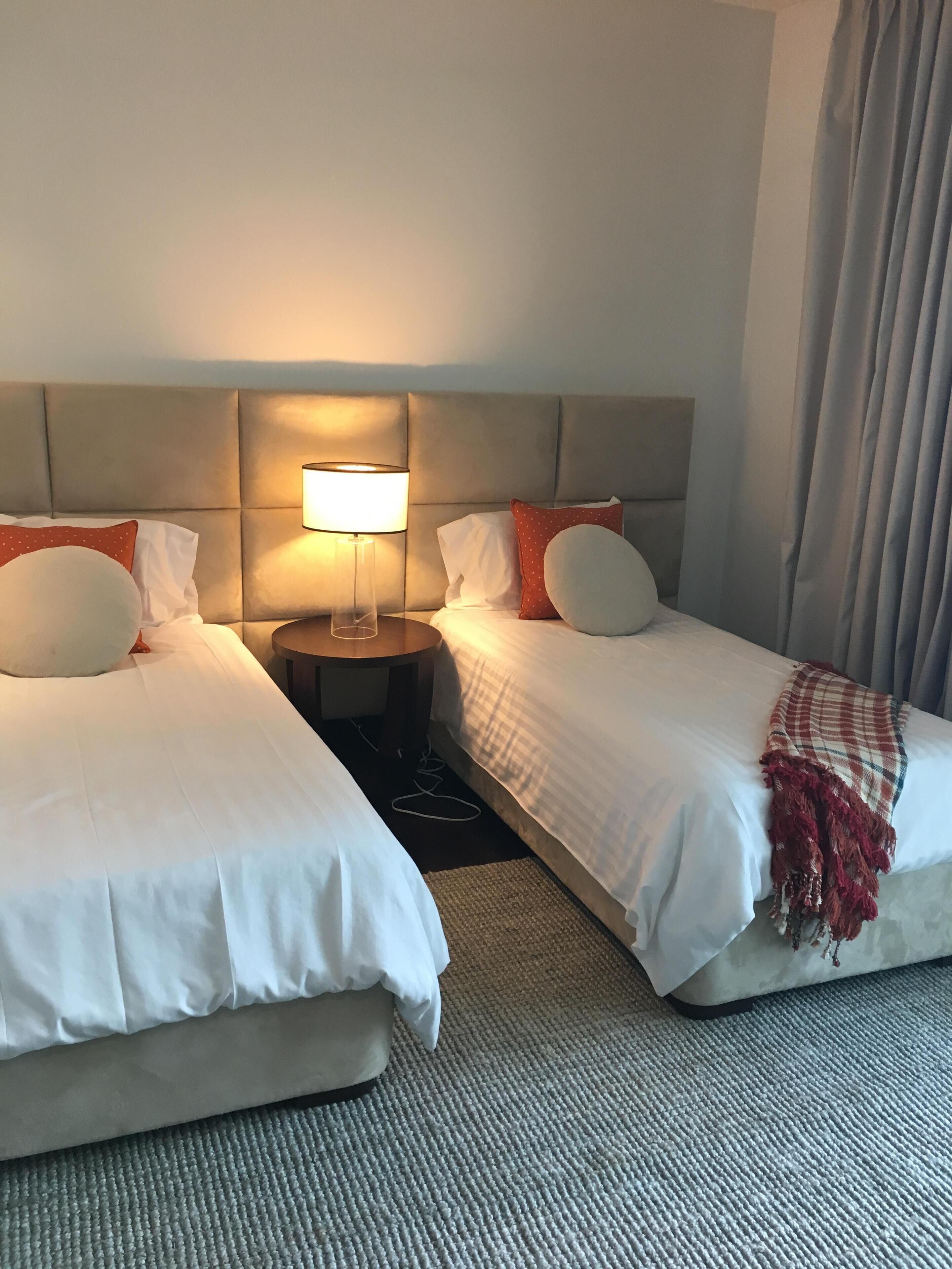 Habitación de hotel con dos camas, una mesita de noche con lámpara y vistas al exterior.