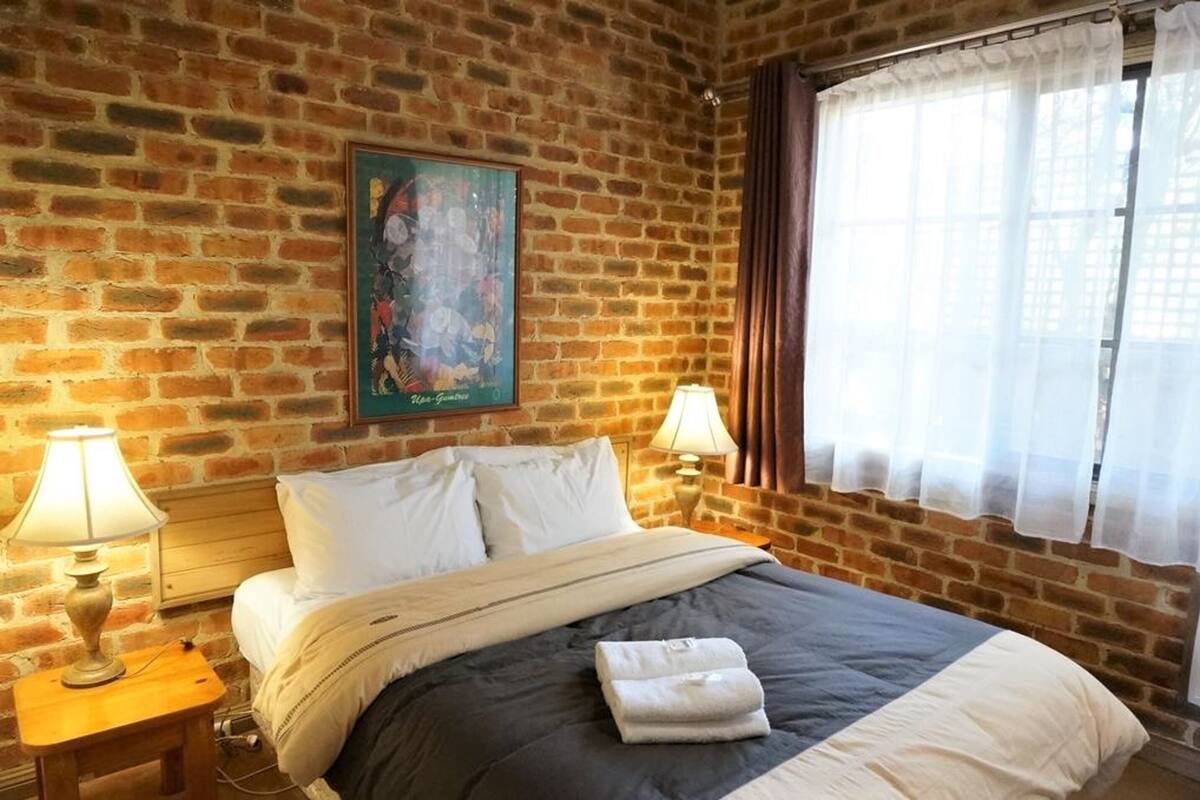 A hotel room with a brick wall, a bed with white bedding, two bedside lamps, and a window with curtains.
