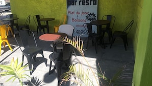 Un espace extérieur aménagé avec des tables et des chaises, une enseigne publicitaire pour un restaurant et une vue partielle d’un véhicule stationné.