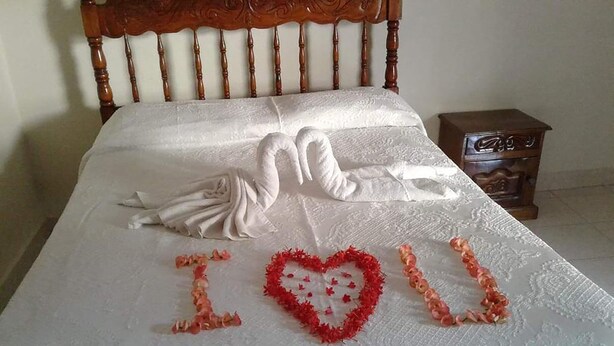 Un lit recouvert d’une courtepointe blanche, orné d’un cœur décoratif composé de fleurs rouges et roses, sur lequel on peut lire les mots « Je t’aime ».