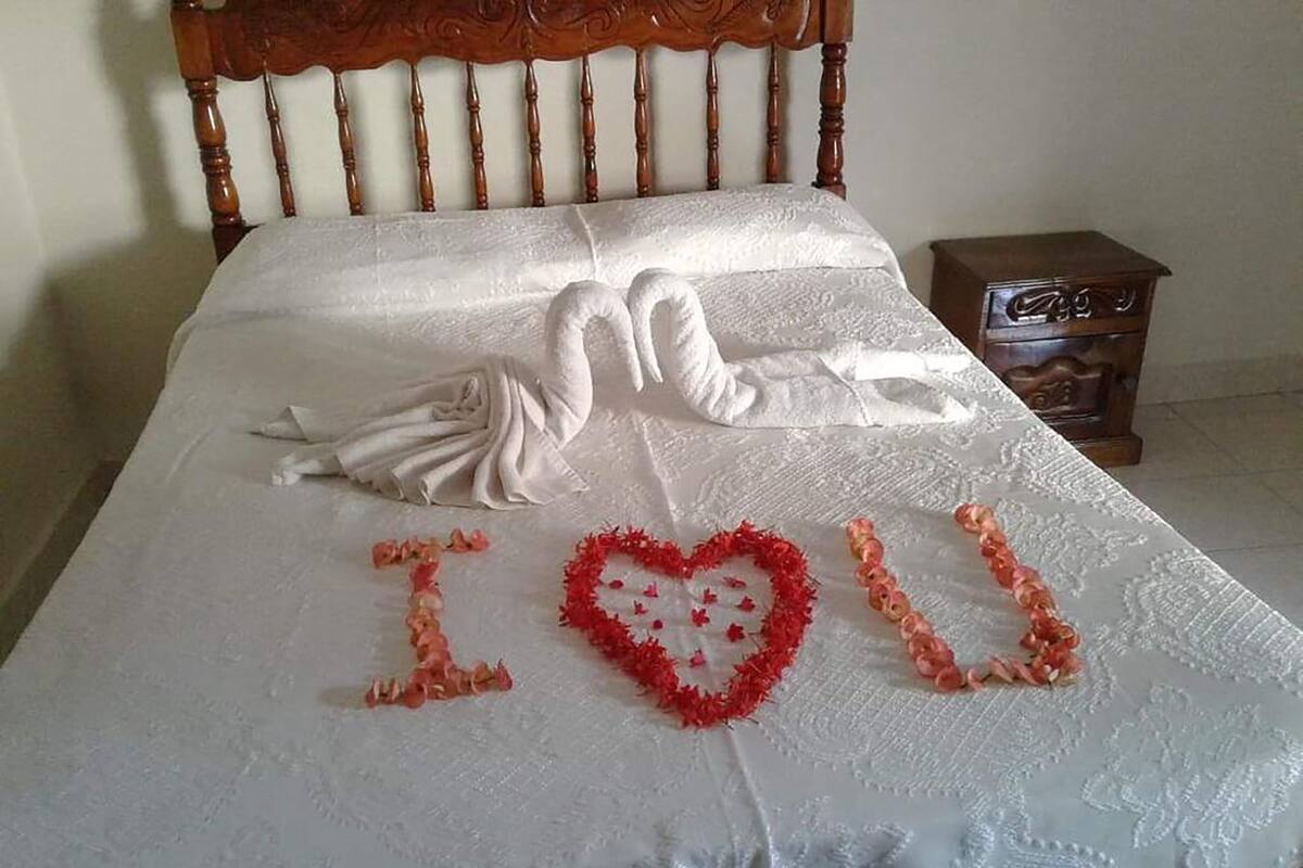 Un lit recouvert d’une courtepointe blanche, orné d’un cœur décoratif composé de fleurs rouges et roses, sur lequel on peut lire les mots « Je t’aime ».