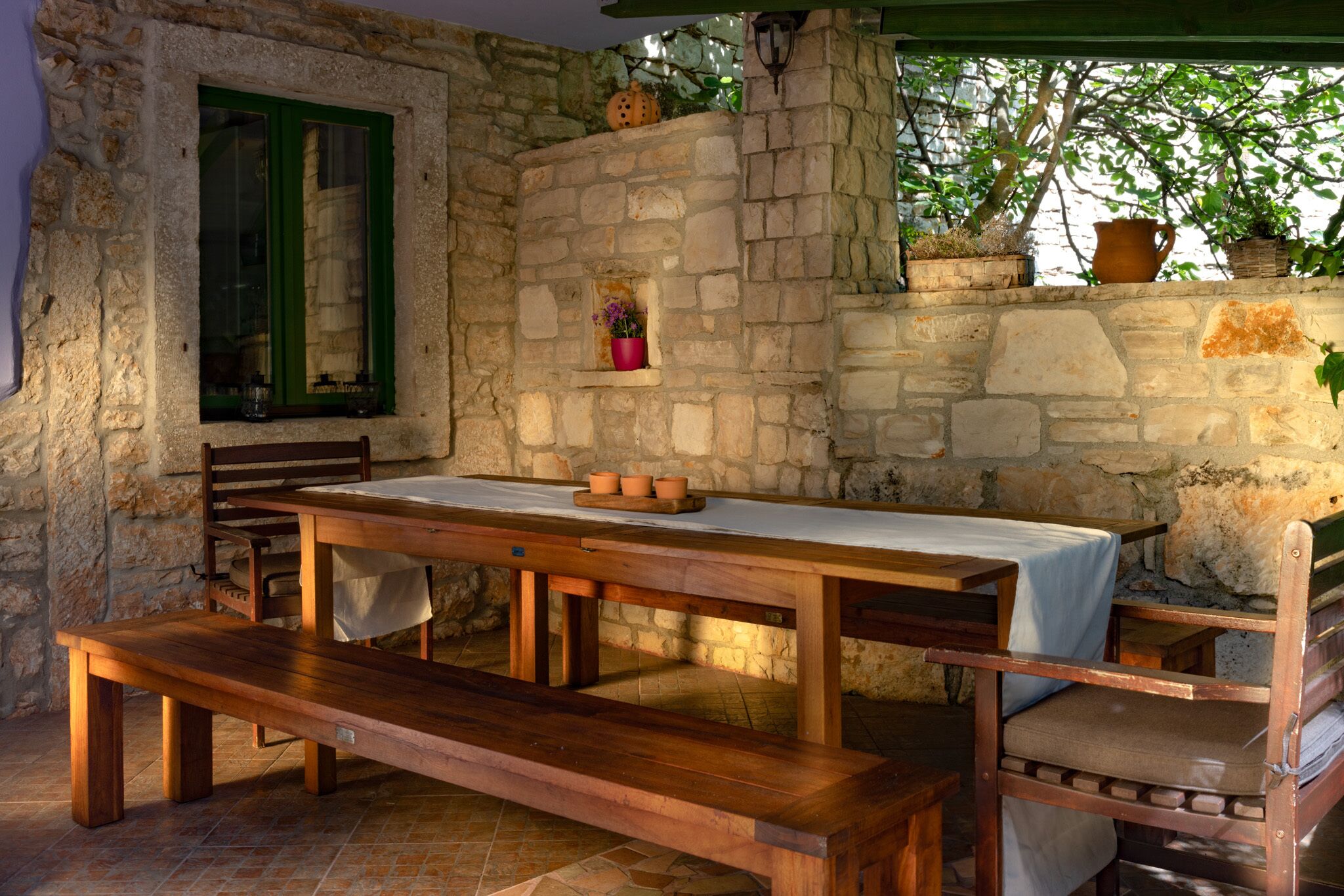 A rustic stone wall with a window, a wooden table with a bench, and potted plants.