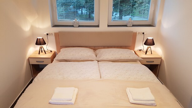 Ein ordentlich bezogenes Bett mit zwei Kissen, ein Nachttisch mit Lampe und ein Fenster mit Jalousien.