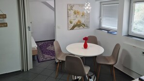 Ein Essbereich mit einem runden Tisch, vier Stühlen, einer roten Vase, einer Deckenleuchte, einem Fenster mit Jalousien und einer Tür zu einem anderen Raum.
