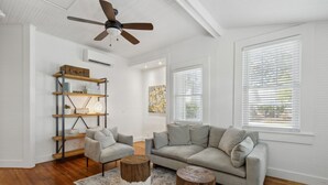 Un soggiorno con un divano grigio, due poltrone, un tavolino in legno e una ventola a soffitto. Si notano scaffali in legno con libri e oggetti decorativi, e un'ampia finestra con tende.