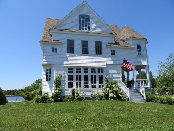 A large white house with a porch, a flag, and a view of a lake. - SALT WATER MAINE COTTAGE WITH DOCK - MINUTES TO BOTANICAL GARDENS (Boothbay)
