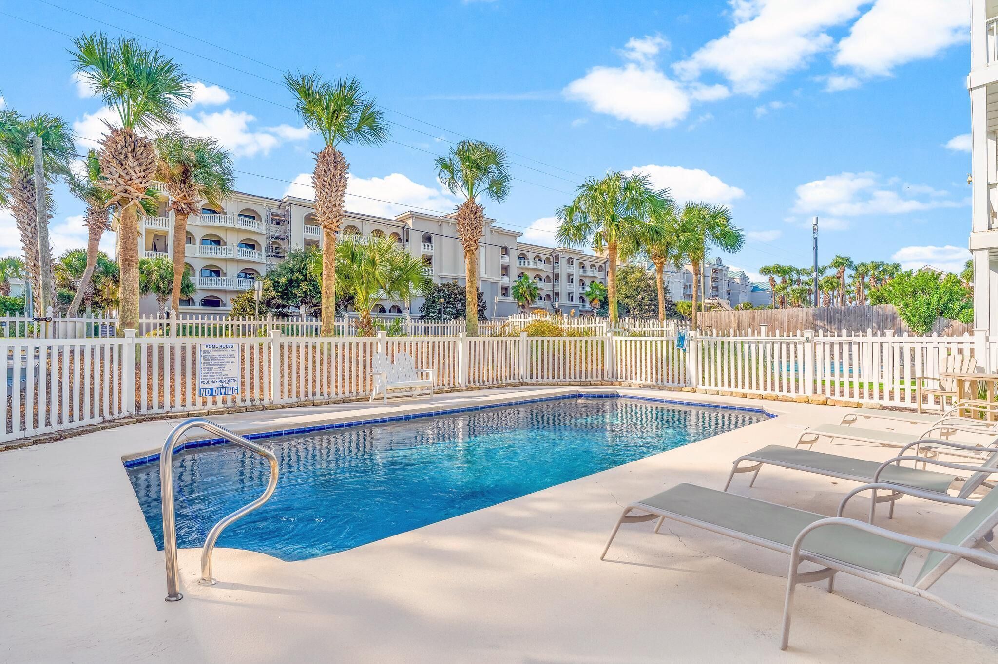 A swimming pool with a white fence, lounge chairs, and palm trees.